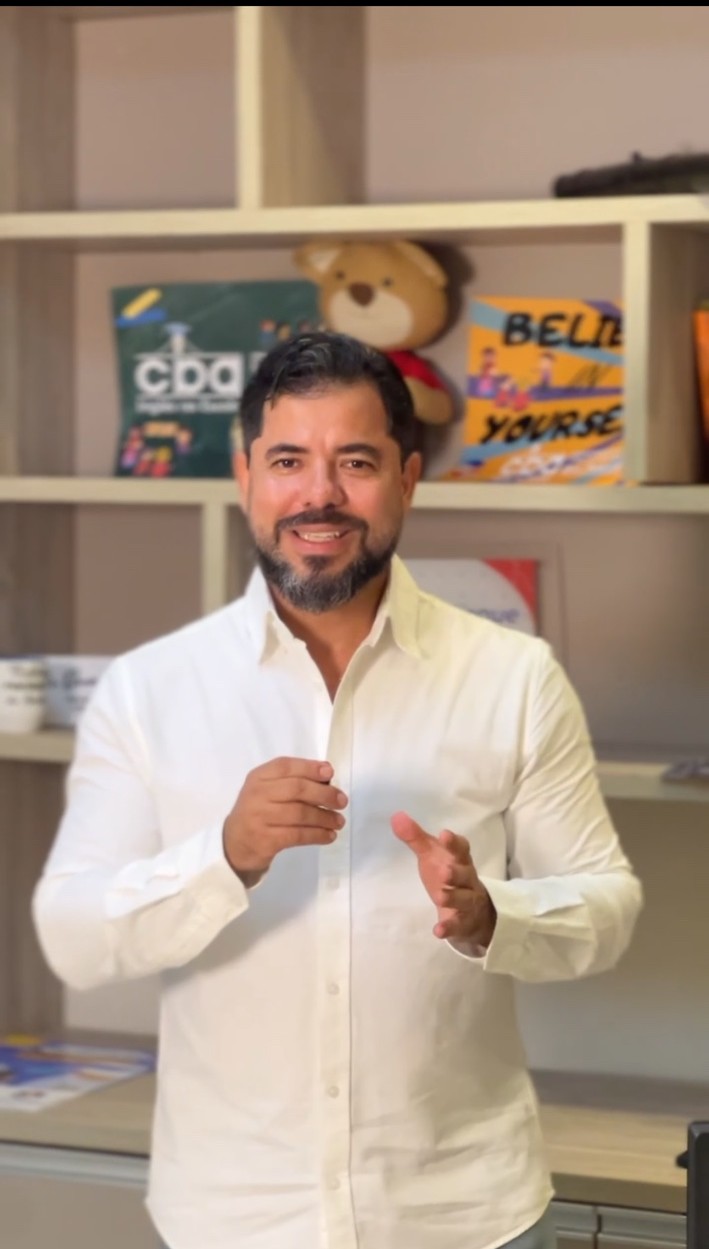Denis Almeida de camisa branca, gesticulando diante de uma estante